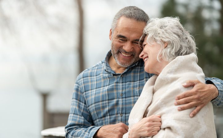 A senior Caucasian woman smiles at her husband of African descent. The couple is standing on a balcony. The man has his arm around the woman as she cuddles in close.